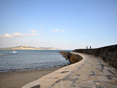 A curved stone walkway along the water with two people walking and boats in the sea at Leys At Valley View Farm in Uplyme