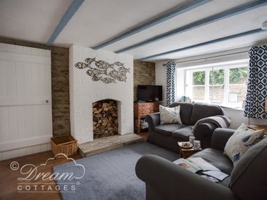 A living room with a stone fireplace, sofas with cushions, a TV on a wooden cabinet and patterned curtains at Lilac Cottage in Burton Bradstock