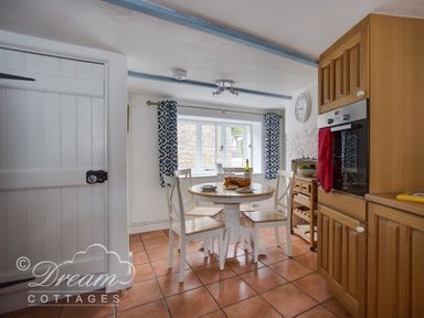 A kitchen with a round table and four chairs near a window with blue patterned curtains at Lilac Cottage Burton Bradstock