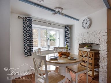 A dining area with a round wooden table and four chairs near a window with patterned curtains at Lilac Cottage in Burton Bradstock