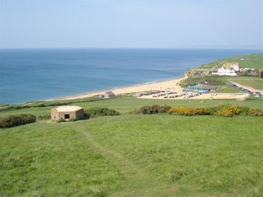A green grassy hill overlooking a beach parking area with cars and buildings near the ocean at Lilac Cottage in Burton Bradstock