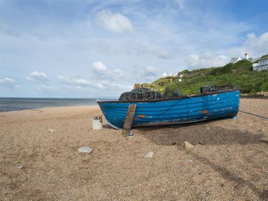 A blue fishing boat on a pebble beach near grassy hills and the sea at Lilac Cottage in Burton Bradstock