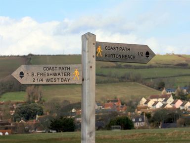 A wooden signpost showing directions to coast path locations with a village and hills in the background at Lilac Cottage in Burton Bradstock