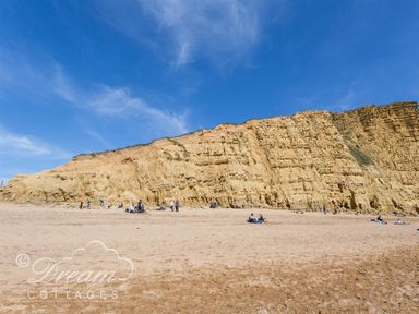 A sandy beach with people sitting near large cliffs under a blue sky at Lilac Cottage in Burton Bradstock