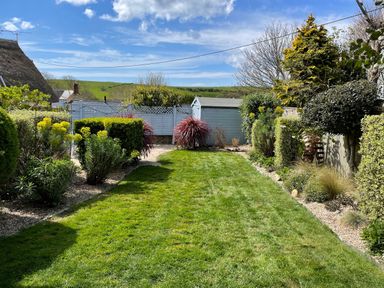 A garden with a lawn surrounded by bushes flowers and a shed at Lilac Cottage in Burton Bradstock