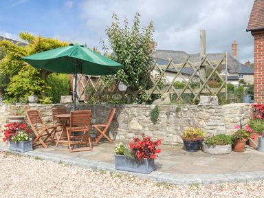An outdoor patio area with wooden table and chairs under a green umbrella surrounded by planters with flowers at Little Forge in West Lulworth
