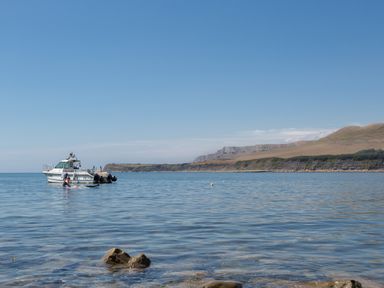 A boat on calm sea with swimmer near rocky shoreline and hills in the background at Little Forge in West Lulworth