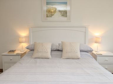 A bed with striped bedding and two pillows with sailboat designs between two white bedside tables with lamps and items on them at Little Forge in West Lulworth