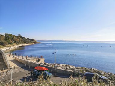 A coastal promenade with people walking and a car with kayaks on the roof near calm water at Lobster Cottage in Brewers Quay Harbour