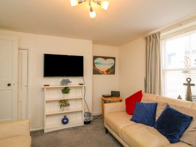 A living room with a mounted television above a white shelf with plants and a vase and a beige sofa with red and blue cushions at Lobster Cottage in Brewers Quay Harbour