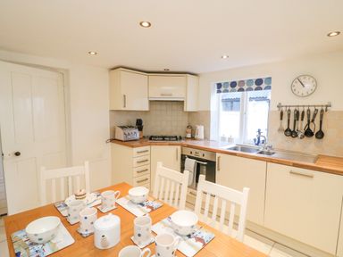 A kitchen with dining table set with mugs bowls and teapot next to countertop with sink stove and hanging utensils at Lobster Cottage in Brewers Quay Harbour