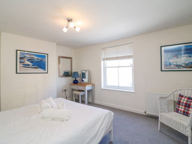 A bedroom with a bed, white wicker chair, small desk with lamp, and framed pictures at Lobster Cottage in Brewers Quay Harbour