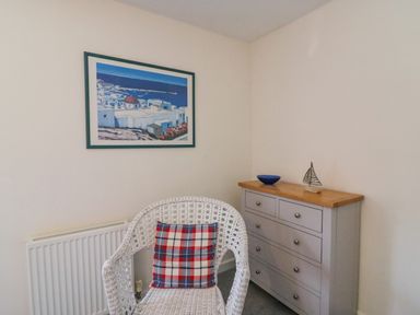 A white wicker chair with a plaid cushion next to a gray dresser with a bowl and sailboat decoration on top at Lobster Cottage in Brewers Quay Harbour