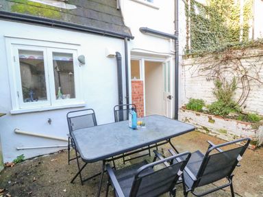A patio with a metal table and four chairs next to a white building with a window and an open door at Lobster Cottage Brewers Quay Harbour