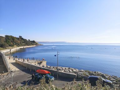 A coastal walkway with people exercising near a calm sea with kayakers and swimmers at Lookout in Cove Castle