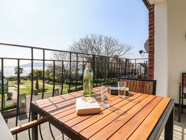 A balcony with a wooden table holding bottles, glasses, and books overlooking a garden and sea at Lookout in Cove Castle