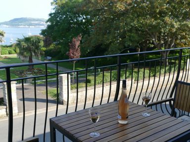 A balcony with a wooden table holding a bottle and two glasses overlooking a garden and water at Lookout in Cove Castle