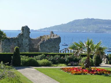 A garden with flowers palm tree stone path and ruins by the sea with boats and hills in background at Lookout in Cove Castle