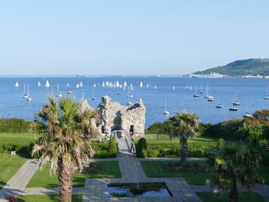 A garden with palm trees and a stone ruin overlooking a sea with many sailboats at Lookout in Cove Castle