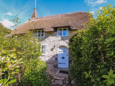 A stone cottage with a thatched roof and a white arched door surrounded by green bushes at Lychgate Cottage in Osmington