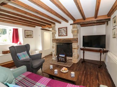 A living room with wooden beams a fireplace an armchair a sofa a wooden coffee table with mugs and cake and a television at Lychgate Cottage in Osmington