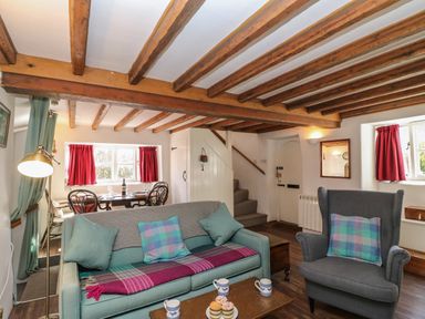 A living room with teal sofa, grey armchair, wooden beams on ceiling, and dining table with red curtains at Lychgate Cottage in Osmington