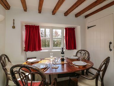A dining area with a wooden table set for four near a window with red curtains at Lychgate Cottage in Osmington