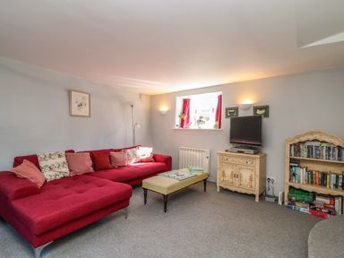 A living room with a red corner sofa a beige ottoman a television on a cabinet and a bookshelf at Lychgate Cottage in Osmington
