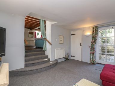 A carpeted living area with a red sofa stairs leading up to another room and glass door with floral curtains at Lychgate Cottage in Osmington