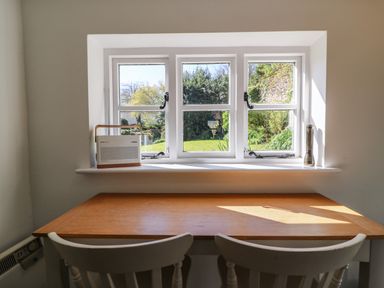 A wooden table with two white chairs by a window with a radio and a flashlight on the windowsill at Lychgate Cottage in Osmington