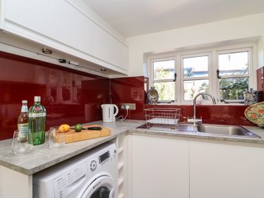 A kitchen countertop with a kettle, bottles, glasses, cutting board, and sink under a window at Lychgate Cottage in Osmington