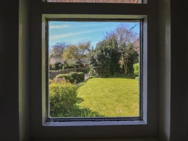 A garden with grass bushes and trees seen through a square window at Lychgate Cottage in Osmington