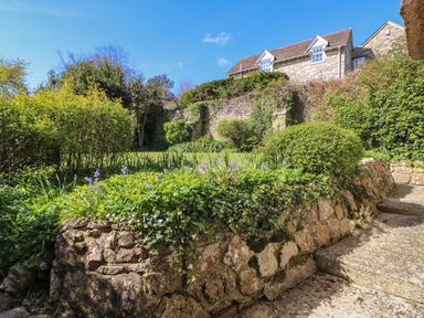 A garden with stone walls shrubs flowers and a stone house in the background at Lychgate Cottage in Osmington