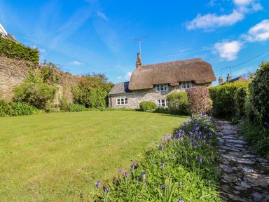 A stone cottage with a thatched roof surrounded by a lawn and garden with flowers and a stone pathway at Lychgate Cottage in Osmington