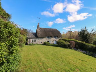 A stone cottage with a thatched roof surrounded by bushes and grass under a blue sky at Lychgate Cottage in Osmington