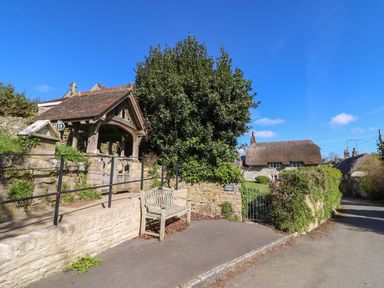 A stone pathway with a wooden bench near a covered structure and a thatched roof cottage in Osmington