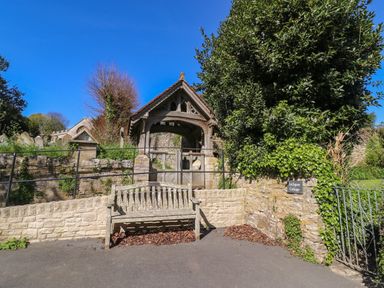 A wooden bench in front of a stone wall and a lychgate with a tiled roof at Lychgate Cottage in Osmington