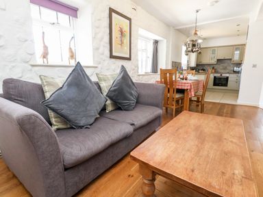 A lounge area with a grey sofa and cushions a wooden coffee table a dining table with four chairs and a kitchen in the background at Magnolia Cottage Osmington