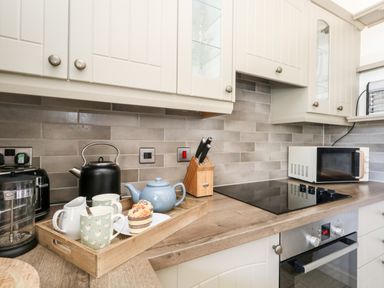A kitchen countertop with a black kettle a blue teapot a wooden tray with mugs a cake a knife block a microwave oven and a built-in stove at Magnolia Cottage Osmington