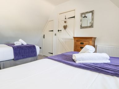 A bedroom with two beds covered with purple blankets and white towels on each bed a wooden chest of drawers a small mirror on the wall and white doors at Magnolia Cottage Osmington in Osmington