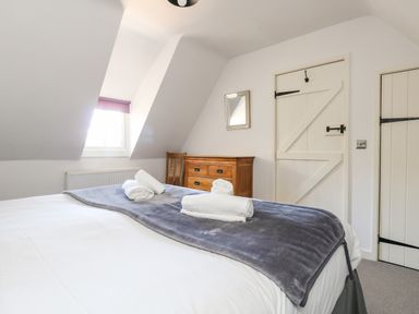 A bedroom with a bed covered with a gray blanket and rolled towels wooden dresser and chair under a window at Magnolia Cottage Osmington
