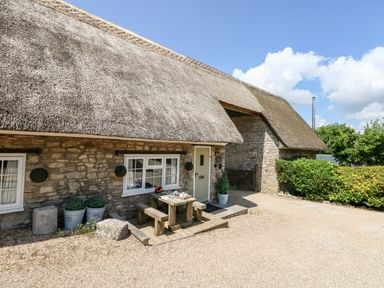 Exterior of a stone cottage with a thatched roof and a wooden picnic table outside at Magnolia Cottage Osmington
