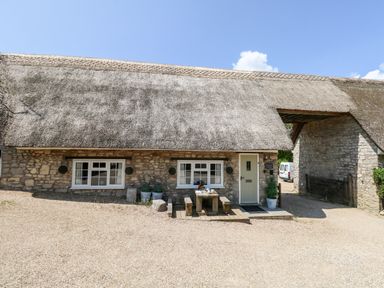 A stone cottage with a thatched roof two windows a wooden door and an outdoor wooden table with benches at Magnolia Cottage Osmington
