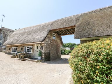 A stone cottage with a thatched roof and outdoor wooden table and benches with shrubbery in front at Magnolia Cottage Osmington