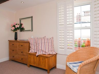 A room with a wooden chest of drawers a wooden storage bench with striped cushions a wicker chair by a window with white shutters at Courtyard House Dartmouth