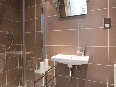 A bathroom with a wall-mounted sink a mirror a glass shower enclosure and a metal stand with toiletries at Courtyard House in Dartmouth