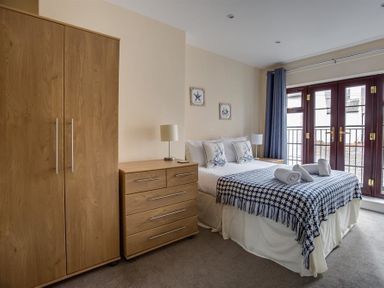 A bedroom with a wooden wardrobe and chest of drawers beside a bed with pillows and folded towels near a window with curtains at New Boathouse in Weymouth