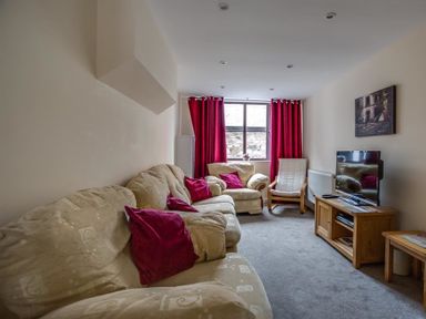 A living room with beige sofas red cushions and curtains a window a TV on a wooden stand and a chair at New Boathouse in Weymouth