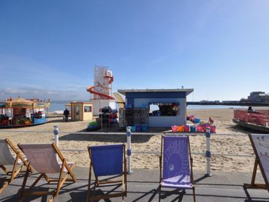A sandy beach with deck chairs in front of a kiosk and amusement rides at New Boathouse in Weymouth