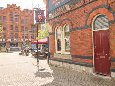 An outdoor seating area with wooden benches and tables near a red brick building with arched windows and a red door at Brewers Quay in Weymouth